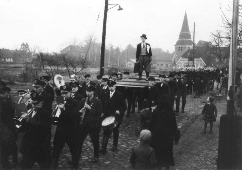 Die Mannschaften der Wasserkunst Curslack und der FF Curslack marschieren zum Curslacker Sportplatz im Frühjahr 1934. Das helle Gebäude links neben dem getragenen Mann mit dem Hut: Das erste Spritzenhaus der FF Curslack.